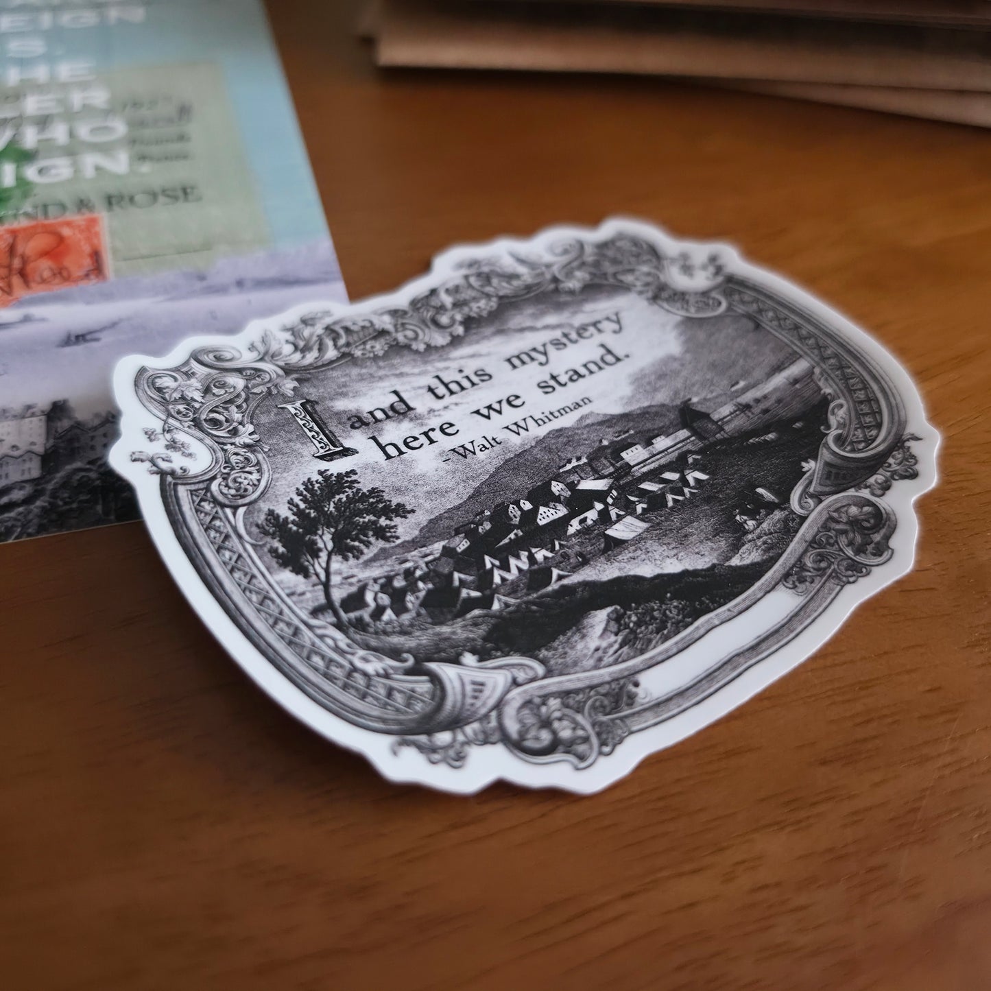 A die-cut sticker featuring a black and white vintage illustration and a quote by Walt Whitman, placed on a wooden tabletop.