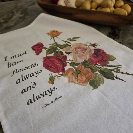 A white tea towel with a printed quote by Claude Monet and a vintage-style rose bouquet, laid on a stone surface with a wooden bowl of lemons in the background.