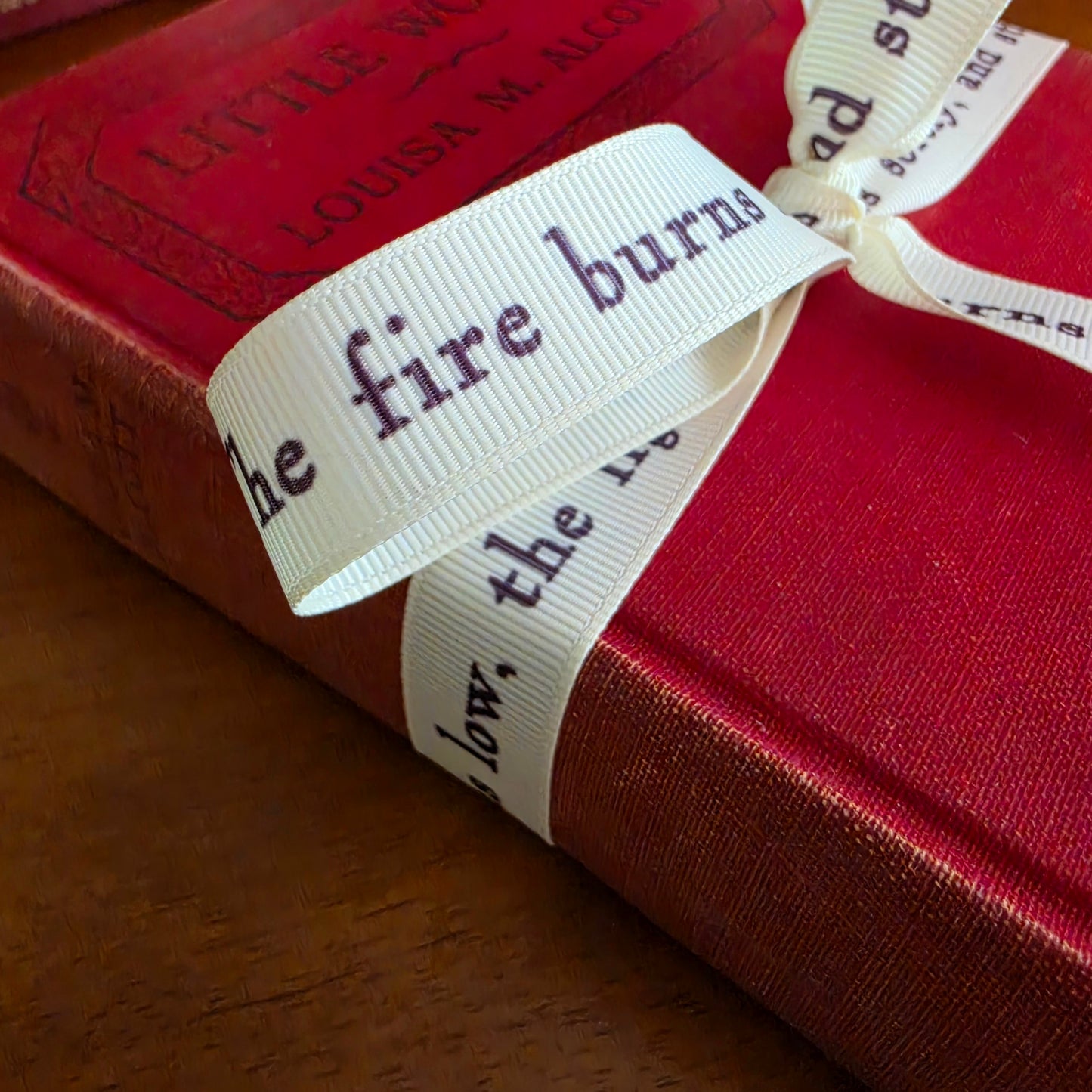 Red book with a white ribbon reading 'The fire burns low, the light falls softly, and still the reader lingers," on a wooden surface.