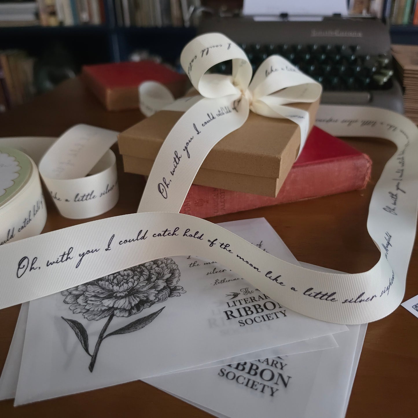 A cream ribbon with elegant black cursive script and a floral line drawing, displayed on a wooden desk with kraft paper gift boxes, a vintage typewriter, and ribbon rolls.