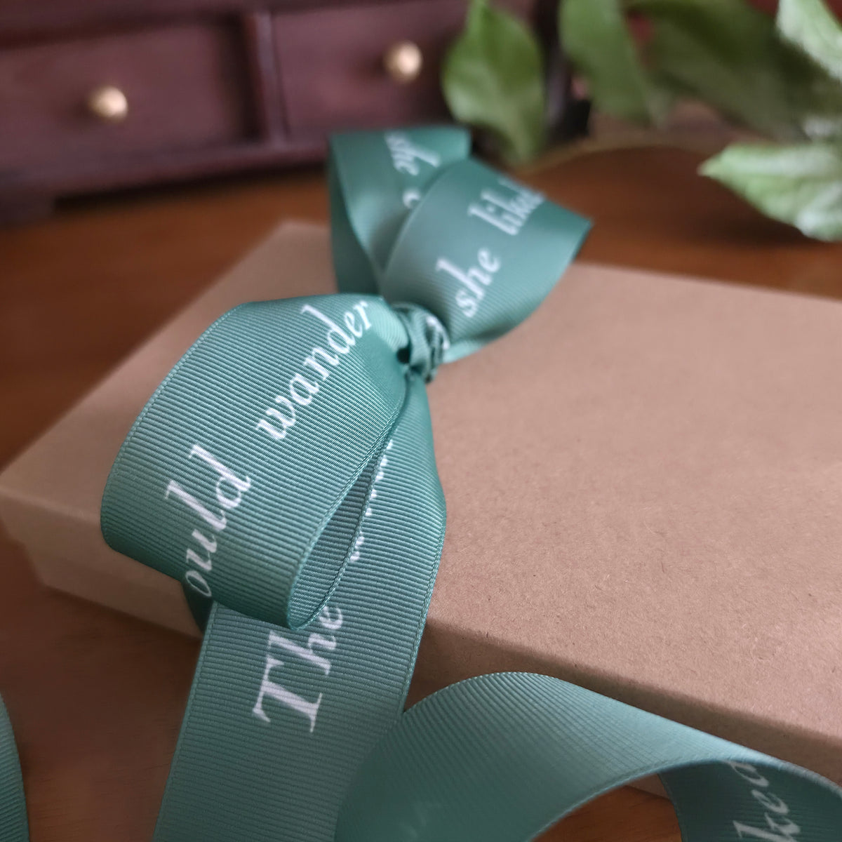 A close-up of a moss green grosgrain ribbon from The Literary Ribbon Collection, with white text and tied in a bow around a brown kraft gift box, set on a wooden surface with a blurred background.