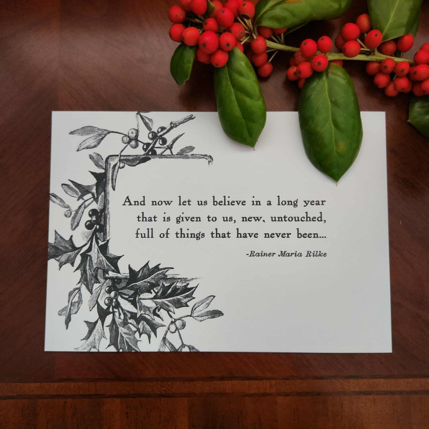 Art print from The Prose & Post Society with holly design and quote on a wooden surface with holly berries.
