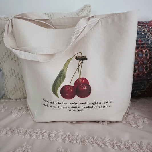A cream canvas tote bag with a printed illustration of two red cherries and a quote from Virginia Woolf, displayed on a textured cream cotton canvas surface with pillows in the background.
