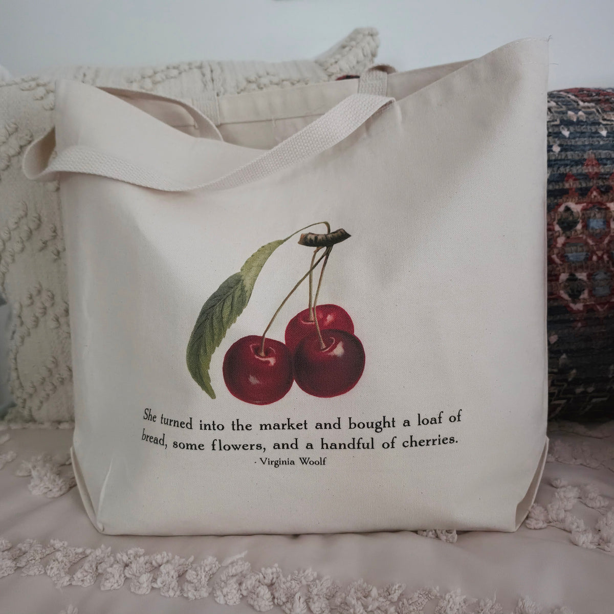 A cream canvas tote bag with a printed illustration of two red cherries and a quote from Virginia Woolf, displayed on a textured cream cotton canvas surface with pillows in the background.