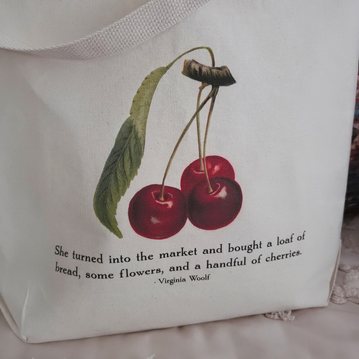 A cream canvas tote bag with a printed illustration of two red cherries and a quote from Virginia Woolf, displayed on a textured cream cotton canvas surface with pillows in the background.