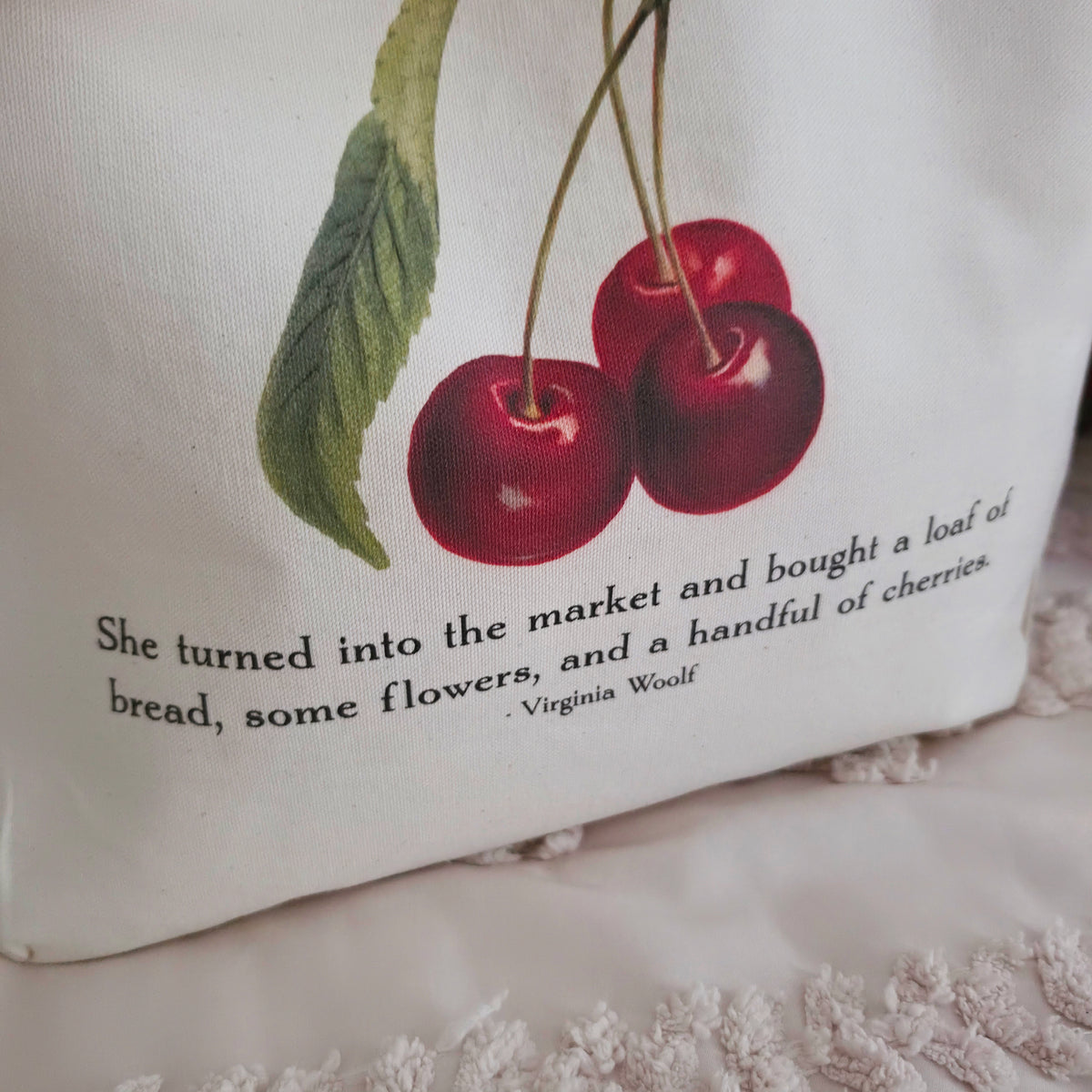 A cream canvas tote bag with a printed illustration of two red cherries and a quote from Virginia Woolf, displayed on a textured cream cotton canvas surface with pillows in the background.