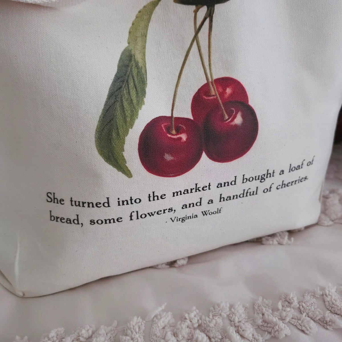 A cream canvas tote bag with a printed illustration of two red cherries and a quote from Virginia Woolf, displayed on a textured cotton canvas surface with pillows in the background.