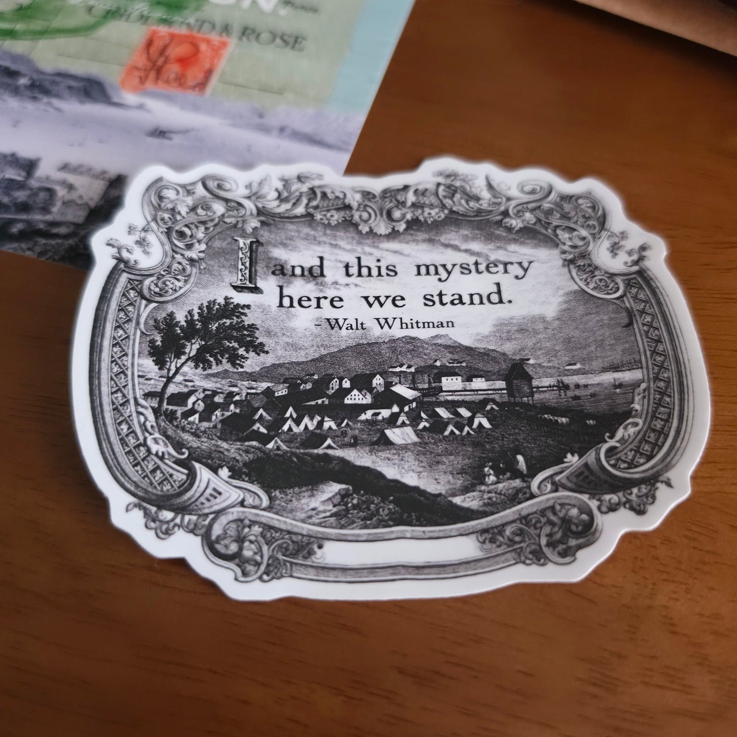 A die-cut sticker featuring a vintage-style black and white illustration with the quote 'and this mystery here we stand. -Walt Whitman' is placed on a wooden tabletop.