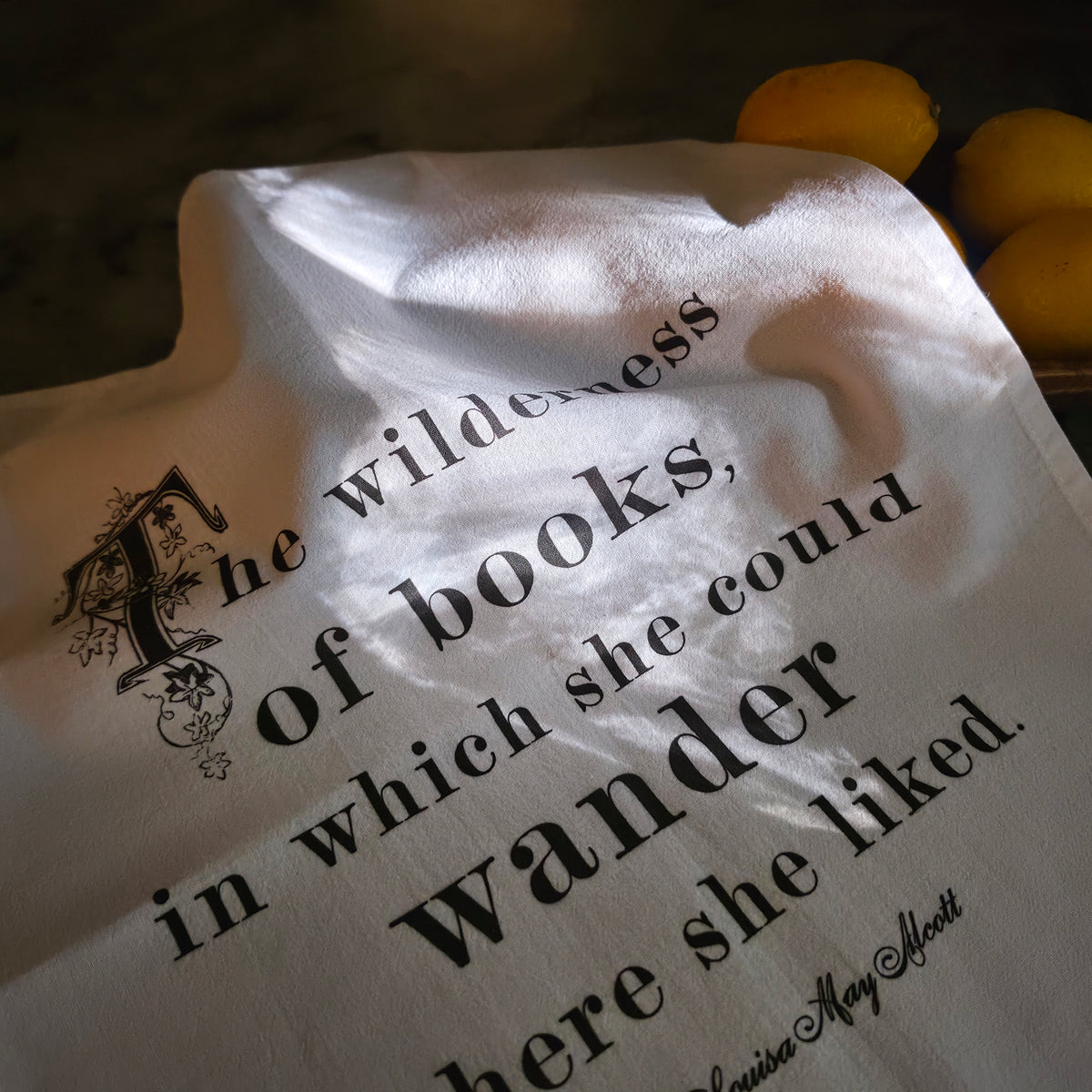 The Wilderness of Books literary tea towel draped in sunlight with Louisa May Alcott quote